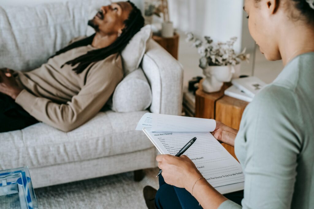 An African man can be seen lying on a sofa and a lady can be seen sitting beside her and writing something on her notepad