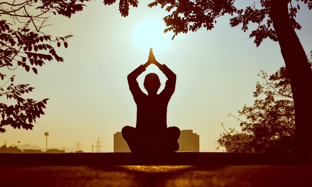 A person is sitting a a yoga pose and meditating with their hands folded and pointed above their head with a clean and serene background of nature