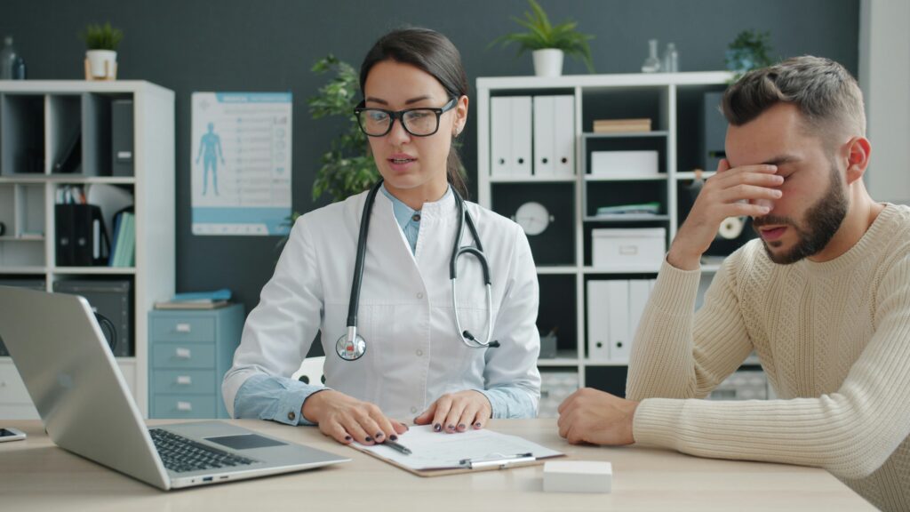 A lady doctor can be seen looking at some reports on her laptop and a man is seen sitting beside her looking worried