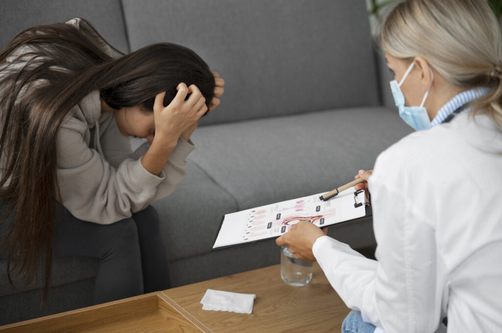 A woman can be seen holding down her head seemingly depressed and another woman who seem like a doctor can be seen sitting beside her and saying something and showing the first woman a piece of paper with something written on it.