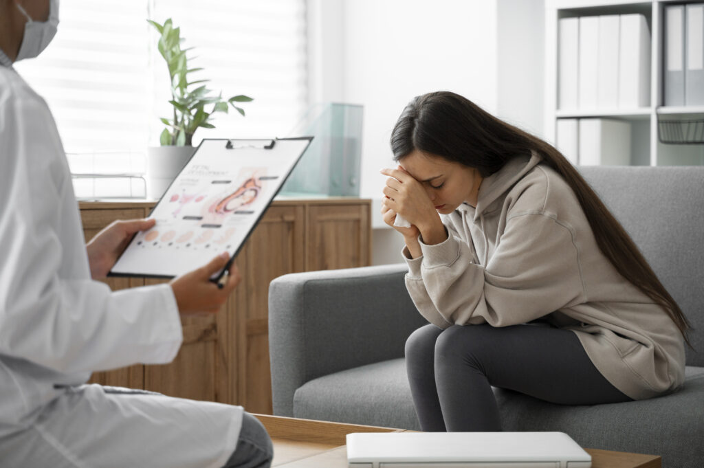 A young woman can be seen sitting on a sofa holding her head with her hands looking down and seemingly sad while a doctor can be seen sitting in front of her holding a report in her hand