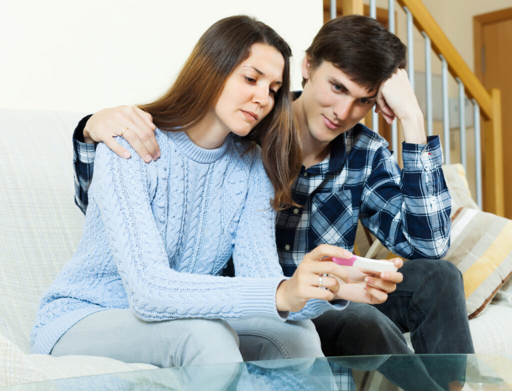 A sad couple can be seen sitting on a sofa and the girl can be seen holding a pregnancy test device