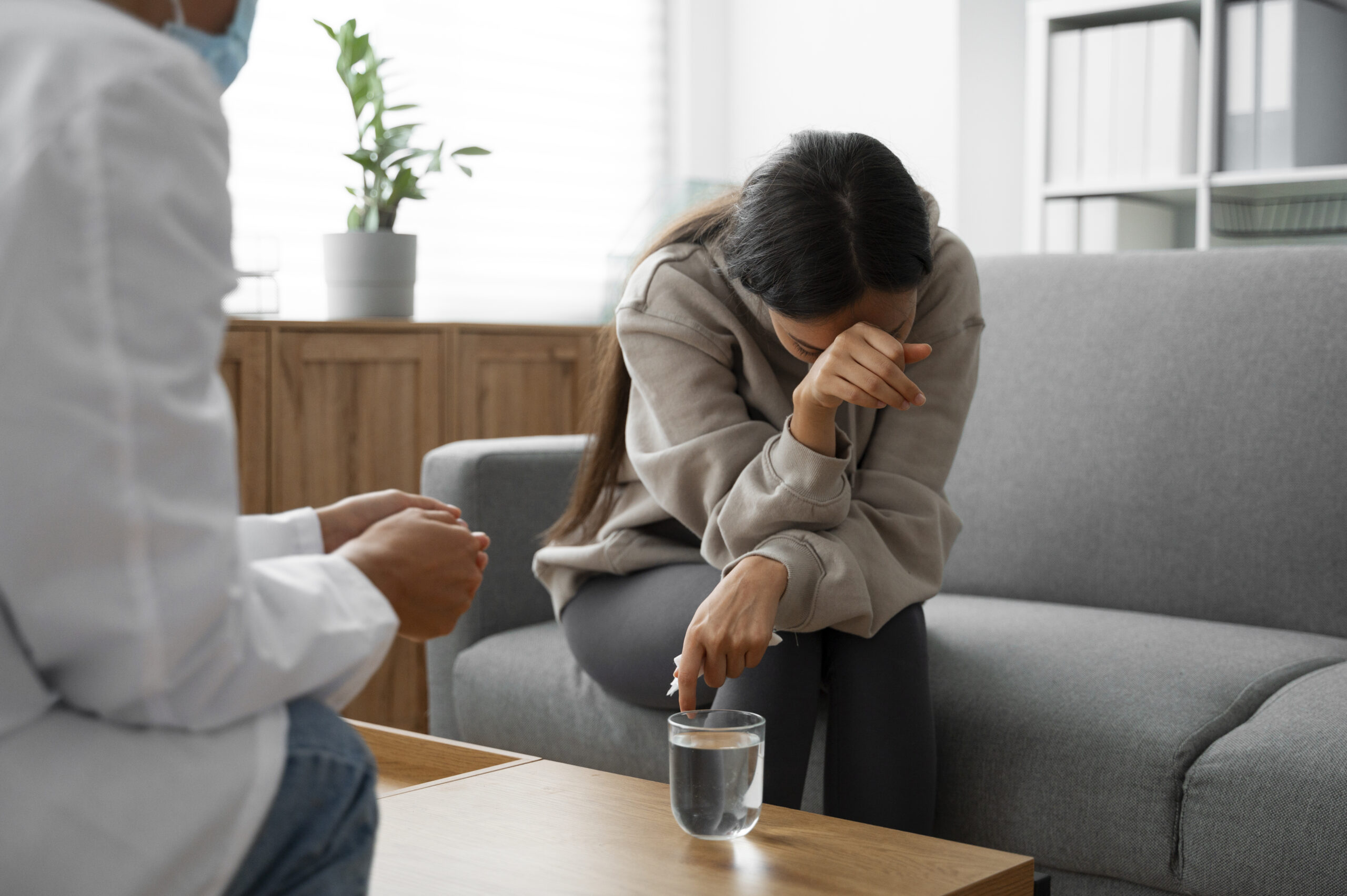 A woman can be seen holding her head down with her hand in a depressed way while a man can be seen sitting in front of her telling her something with a glass of water can be seen on the table in front of her