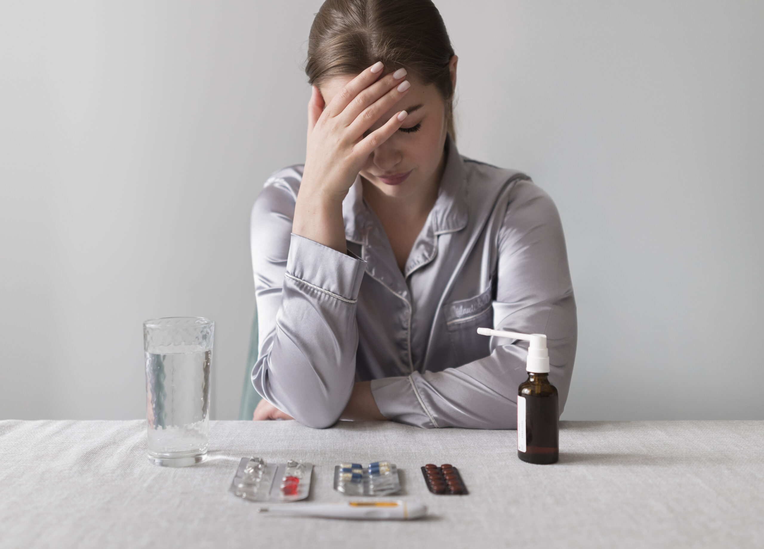 A young woman can be seen holding her head in a tense way with several medicines and a glass of water can be seen lying on a table in front of her