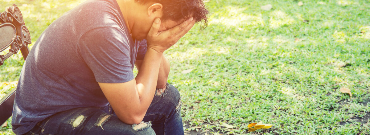 A man can be seen sitting on a bench in a park covering his head in a seemingly tense manner