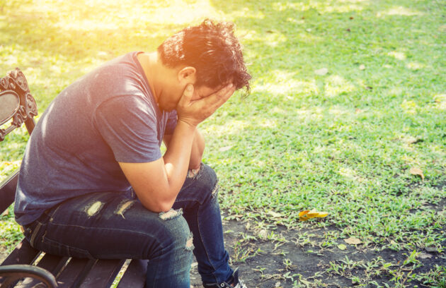 A man can be seen sitting on a bench in a park covering his head in a seemingly tense manner