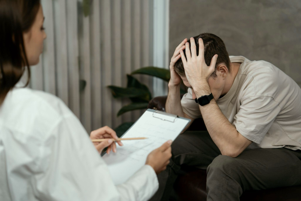 A young man can be seen sitting holding his head in a seemingly frustrating kind of way with a female doctor can be seen sitting in front of him holding a piece of paper and a pen seemingly noting down something