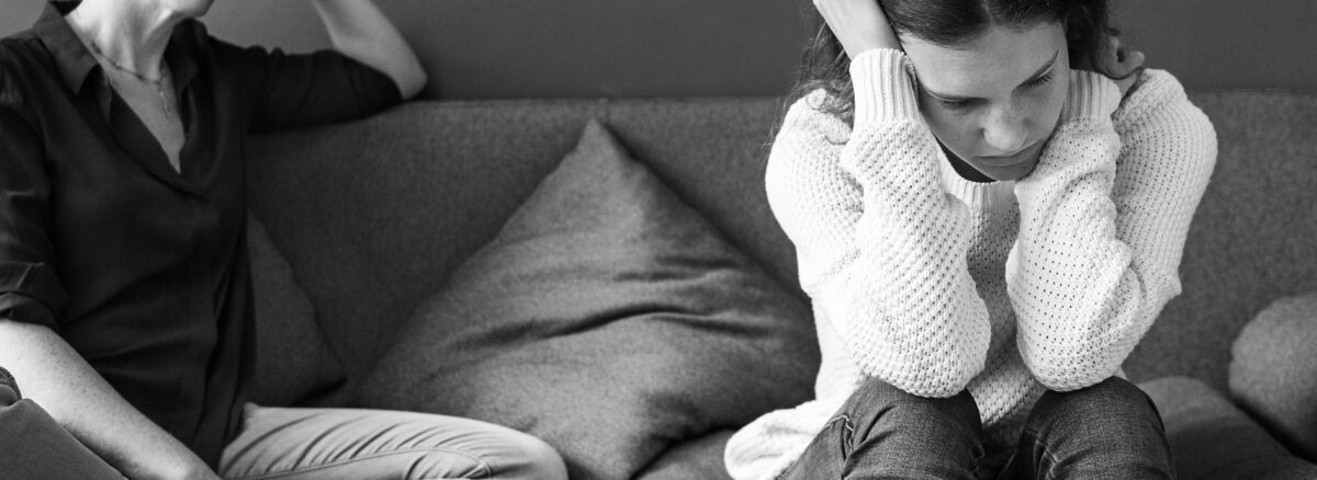 A mother and her teenage daughter can be seen here sitting on a sofa with mother looking at the daughter in an amused way and the daughter seemingly looking pissed and looking away from her