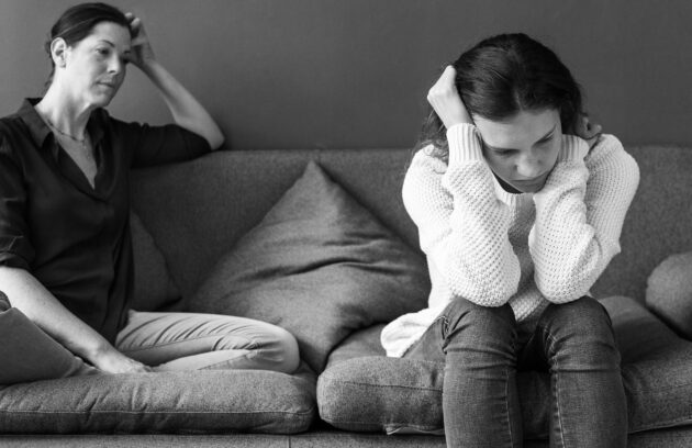 A mother and her teenage daughter can be seen here sitting on a sofa with mother looking at the daughter in an amused way and the daughter seemingly looking pissed and looking away from her