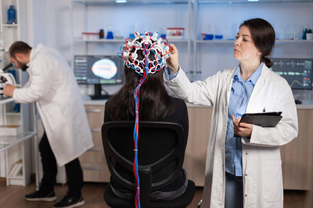 A woman doctor can be seen putting a piece of medical headgear on another woman with lots of wire attached to it and a male doctor can be seen doing something in the background