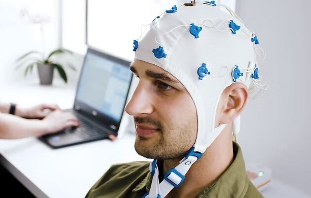 A man can be seen wearing QEEG headset while someone in the background is doing something on their laptop