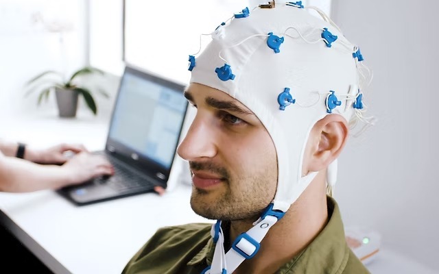 A man can be seen wearing QEEG headset while someone in the background is doing something on their laptop