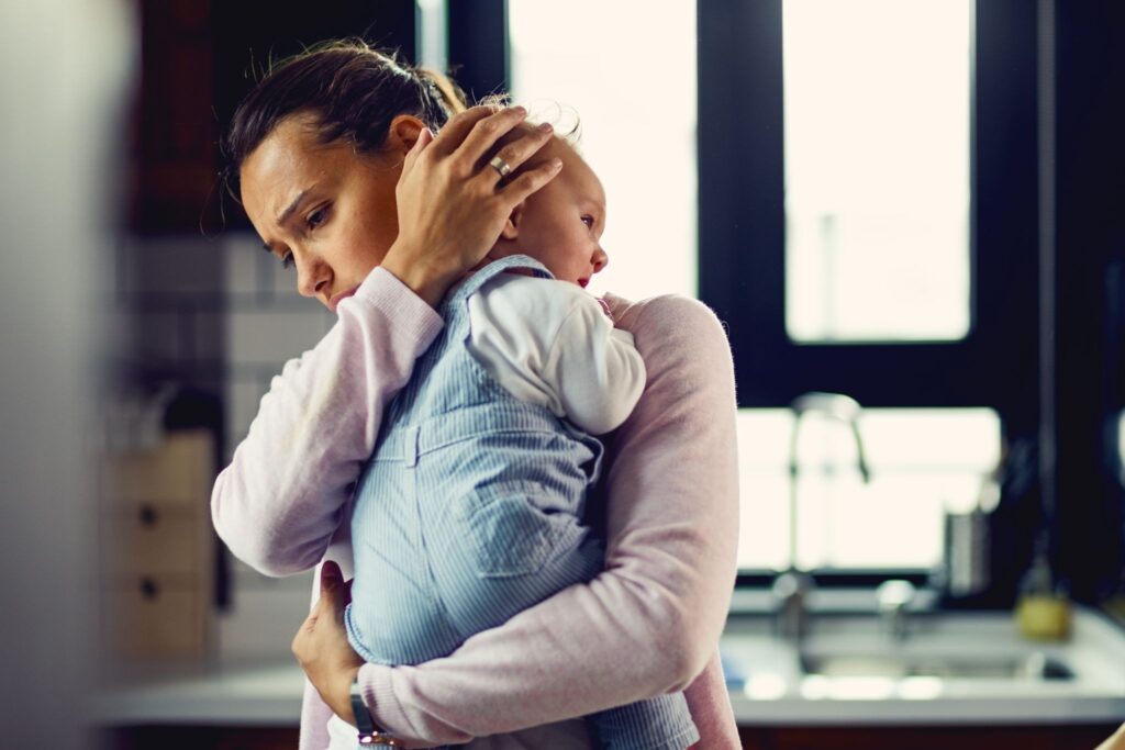 A young mother can be seen holding her baby and seen standing in her kitchen with a blurred background looking tense